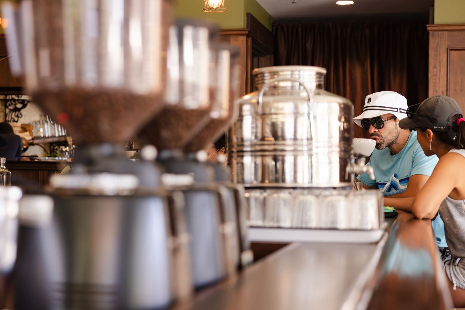 Baristas in blue shirts work behind a coffee shop counter with equipment and metal containers visible in the background.