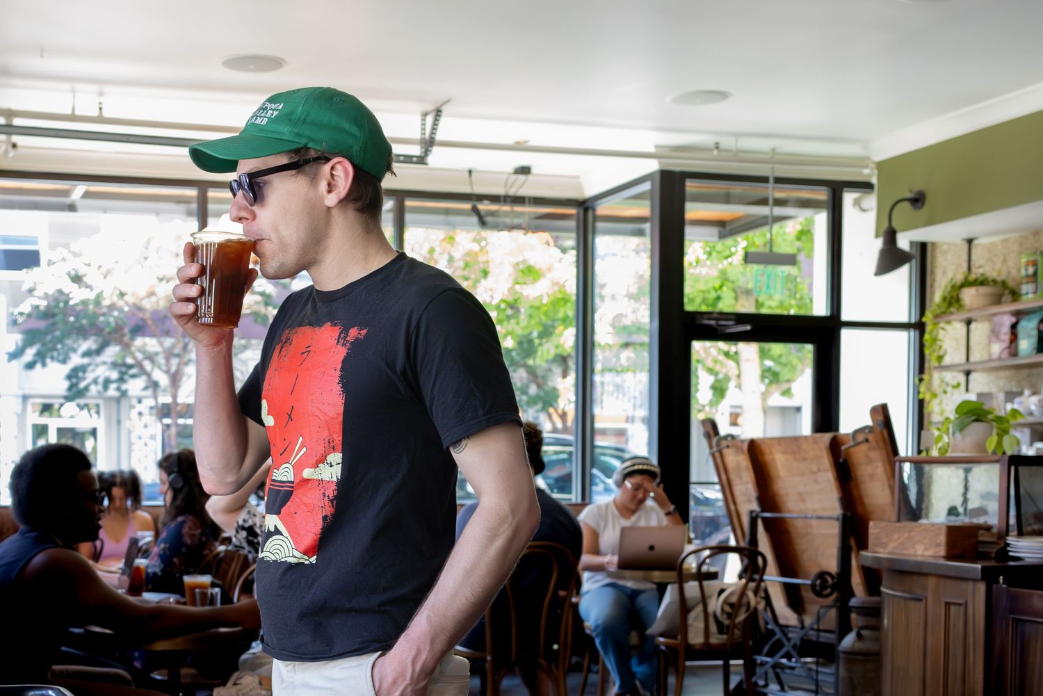Someone in a green baseball cap and black t-shirt drinks coffee while standing in a casual cafe setting.