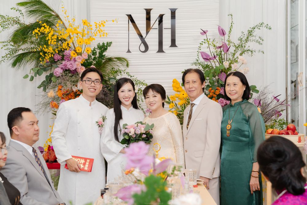 Vietnamese wedding couple poses with family members before monogram backdrop with flowers.