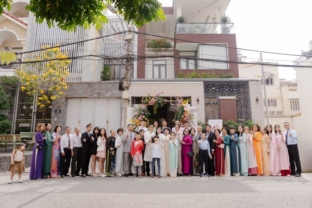 Large Vietnamese wedding party group photo outside multi-story home with floral entrance arch.