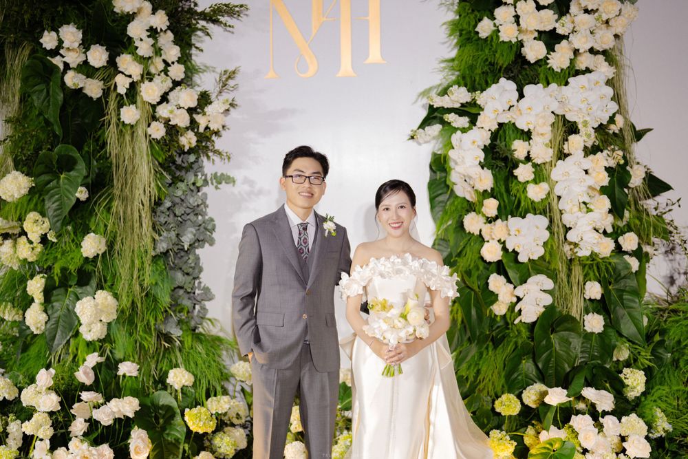 Elegant wedding couple poses before lush green floral backdrop with white rose arrangements.