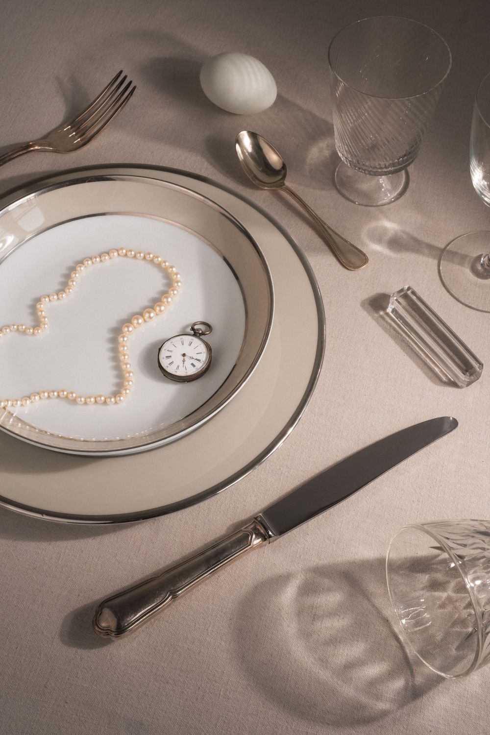 Elegant place setting with pearl heart, pocket watch, and crystal glasses on white tablecloth.