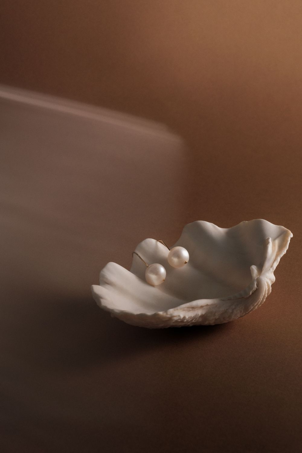 White seashell dish holding two elegant pearls against a moody brown background.