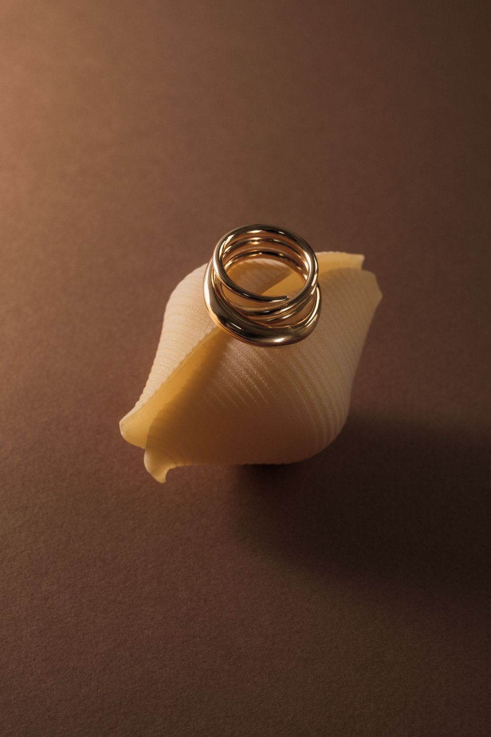 Gold spiral ring balanced on a curved pasta shell against brown backdrop.