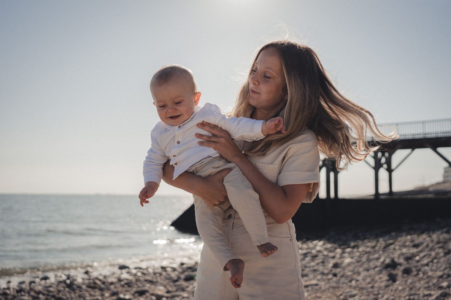 Parent tenant son bébé sur la plage, avec les vagues et une jetée en arrière-plan.