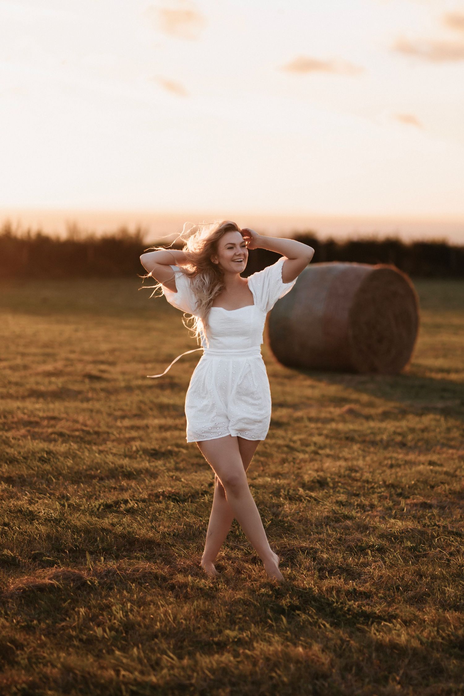 A person in a white summer dress dances joyfully in a golden field at sunset with a hay bale in the background.