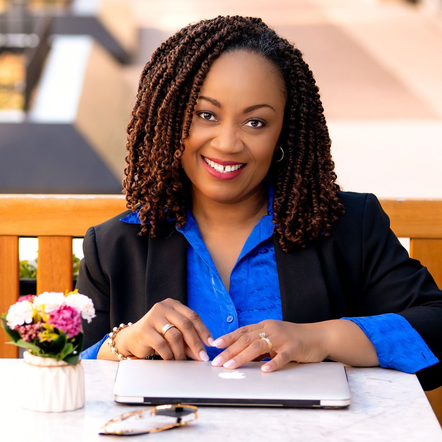 Professional in a black blazer and blue shirt working at a desk with a laptop and flowers nearby.