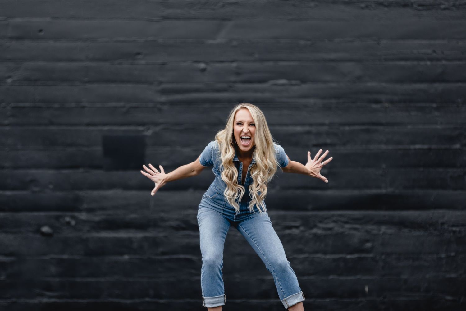 Energetic woman with wavy blonde hair wearing jeans strikes playful pose against dark gray wall.