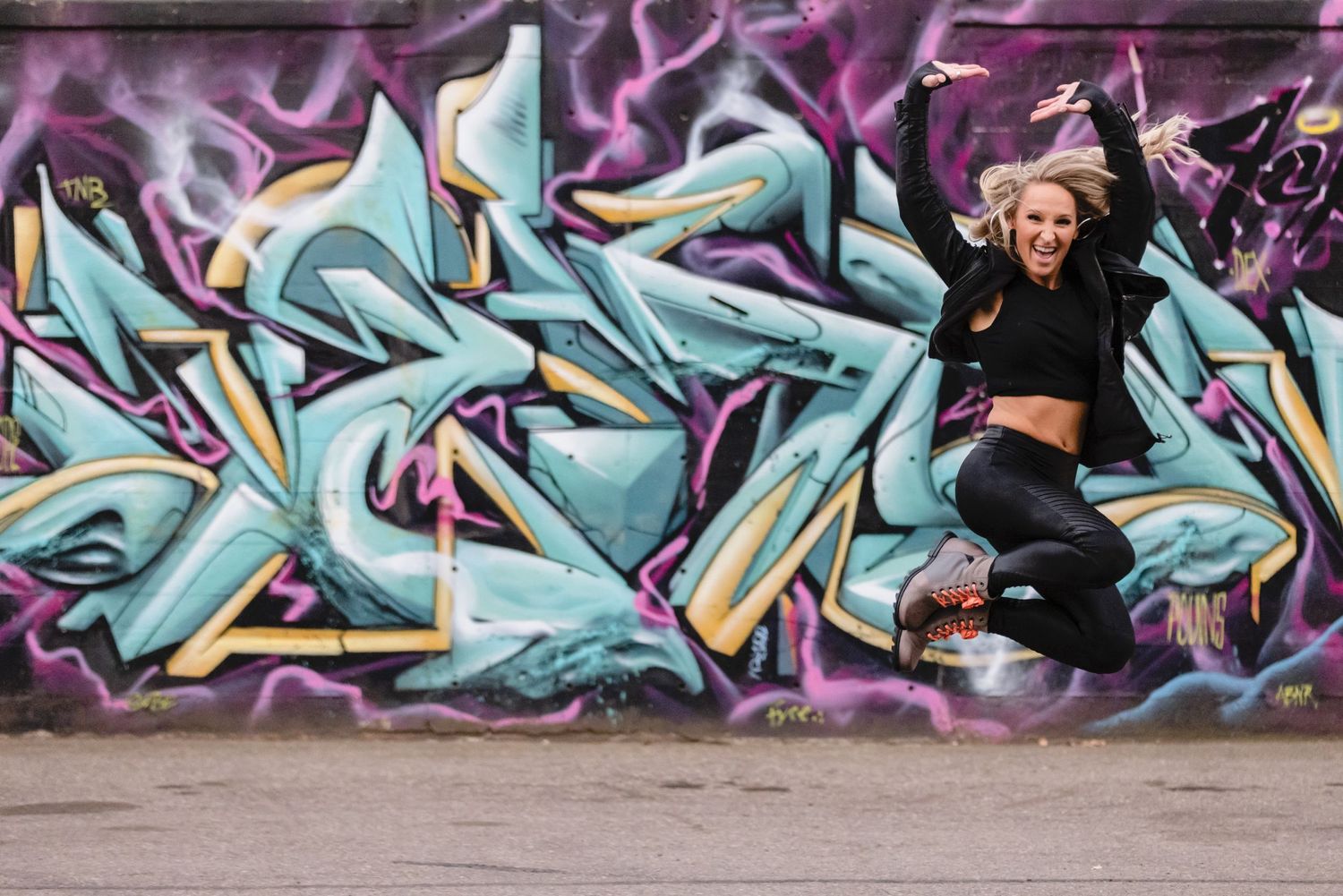 An enthusiastic dancer leaps joyfully against a vibrant turquoise and purple graffiti wall background.