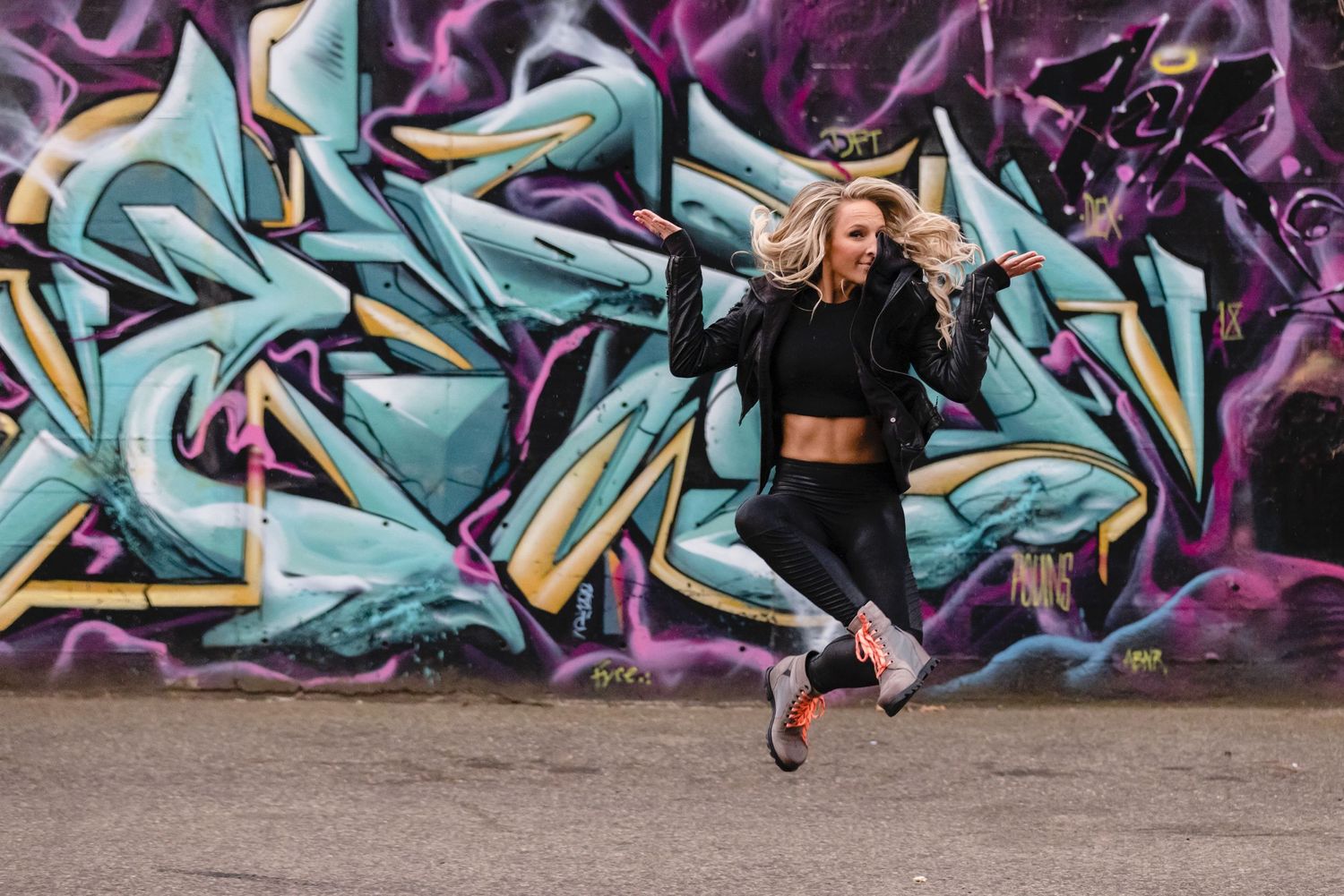 Energetic dancer leaps in front of vibrant turquoise and purple graffiti wall wearing black athletic wear.
