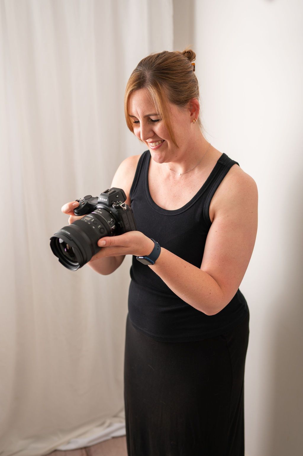 A red-haired photographer in black holds a camera while standing before a lit vanity mirror in a studio.