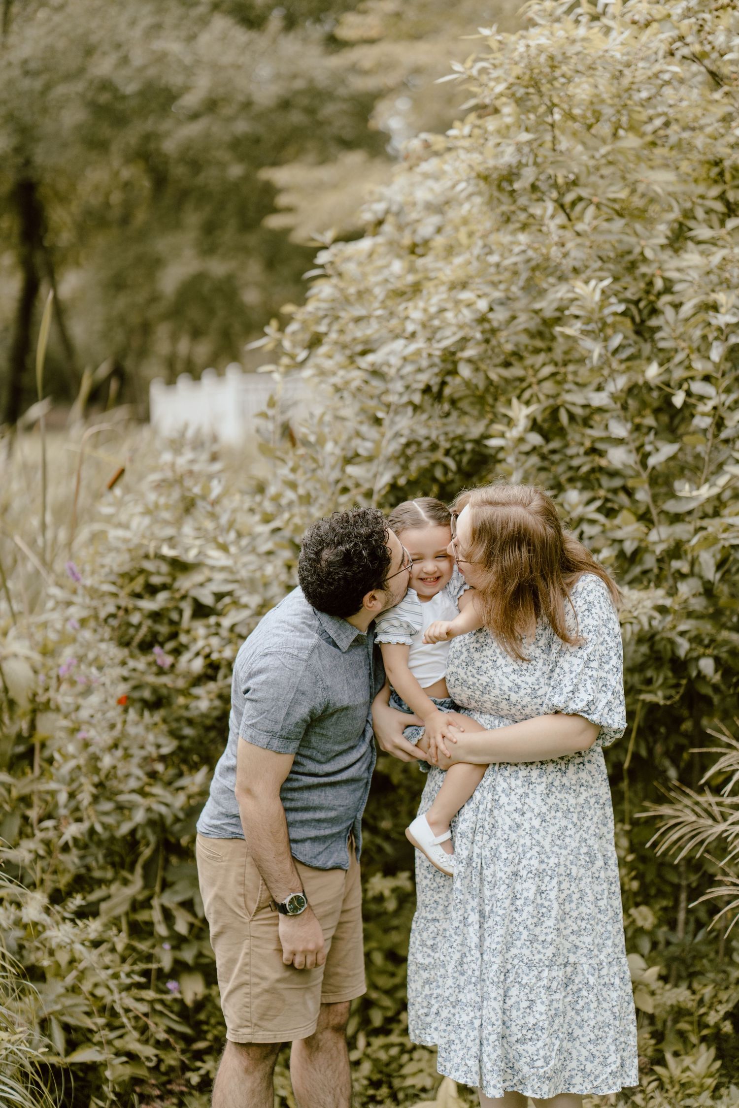A couple and their young child share a tender moment together in a lush garden setting with white flowers.