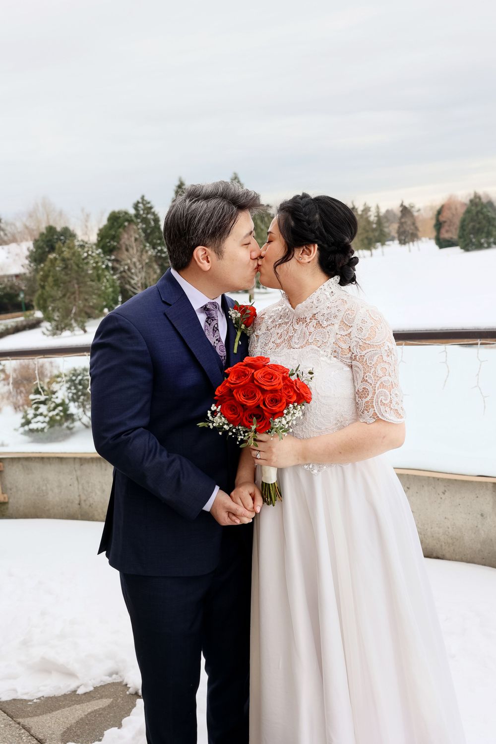 A winter wedding couple share a romantic moment on a snow-covered terrace with a bright red rose bouquet. smilein wedding photography