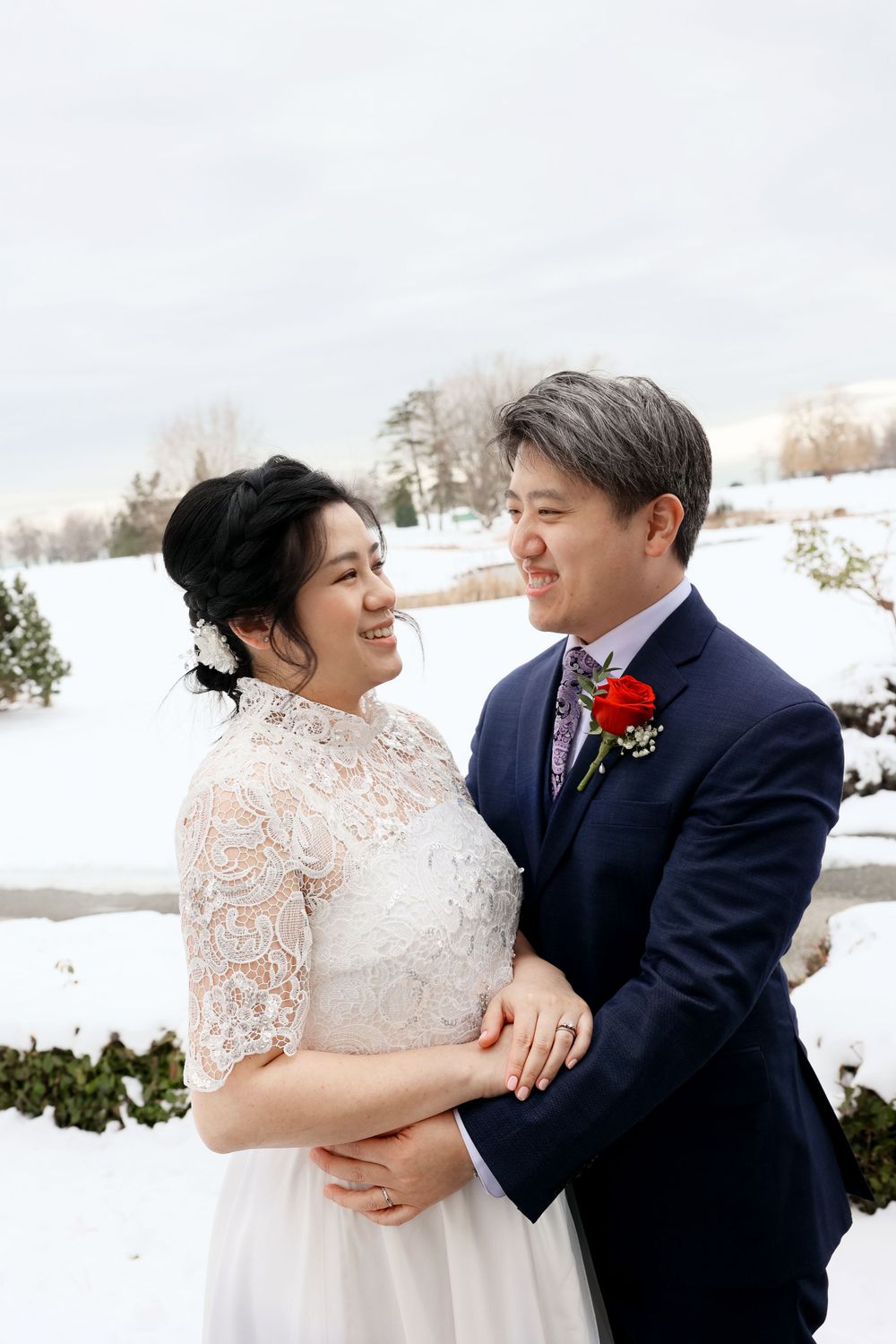 Happy couple embraces in snowy winter wedding scene with lace dress and navy suit in outdoor setting. smilein wedding photography vancouver
