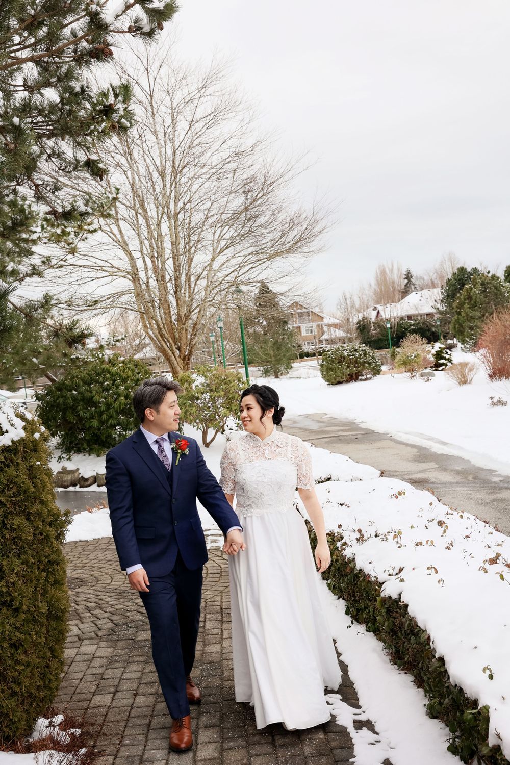 A couple in wedding attire walks together on a snowy winter path bordered by evergreens and bare trees.smilein wedding photography vancouver