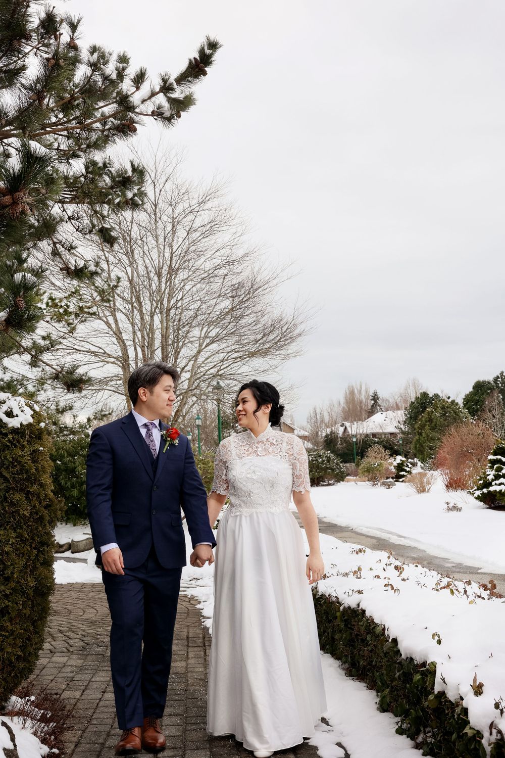 A couple in wedding attire walk together on a snowy winter path with bare trees and evergreens . smilein wedding photography vancouver