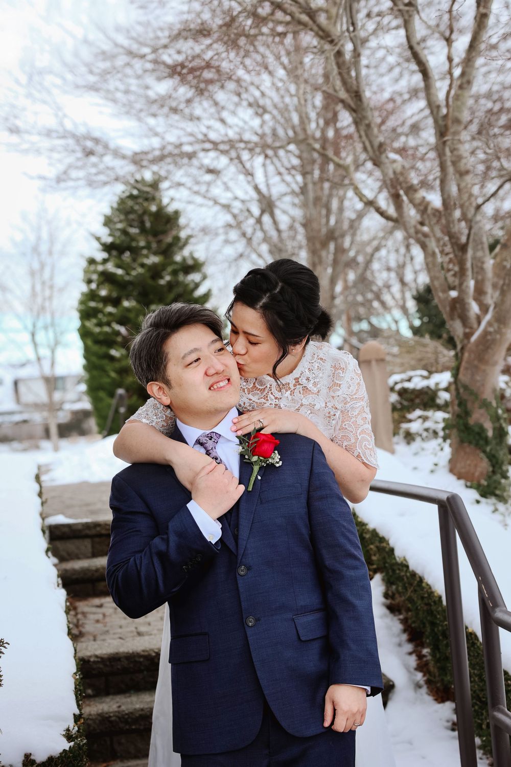 A couple shares a tender moment on a snowy winter day, with the groom wearing a navy suit with red boutonniere. smilein wedding photography