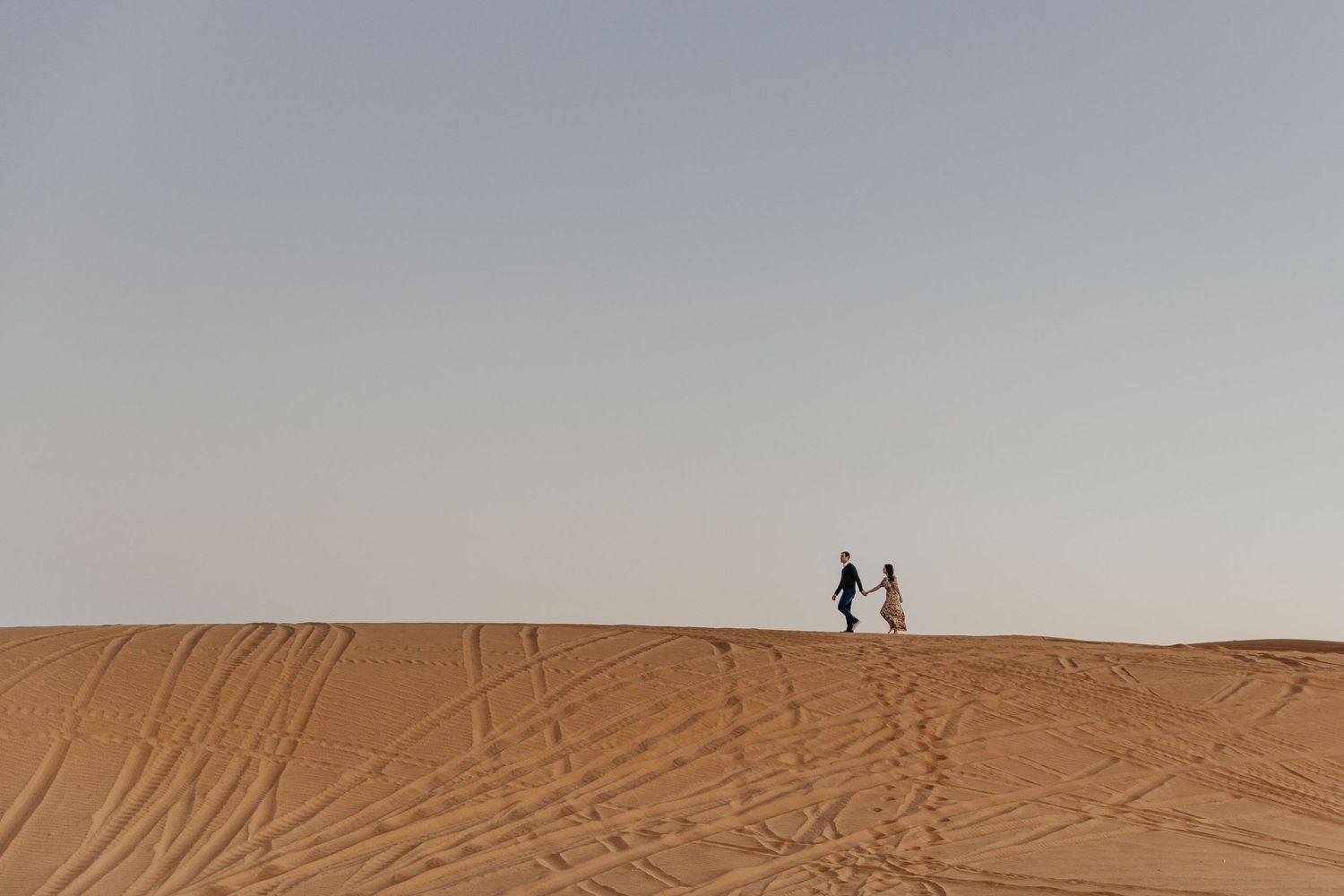 Two silhouetted figures stand atop a sand dune against a hazy sky in a desert landscape.