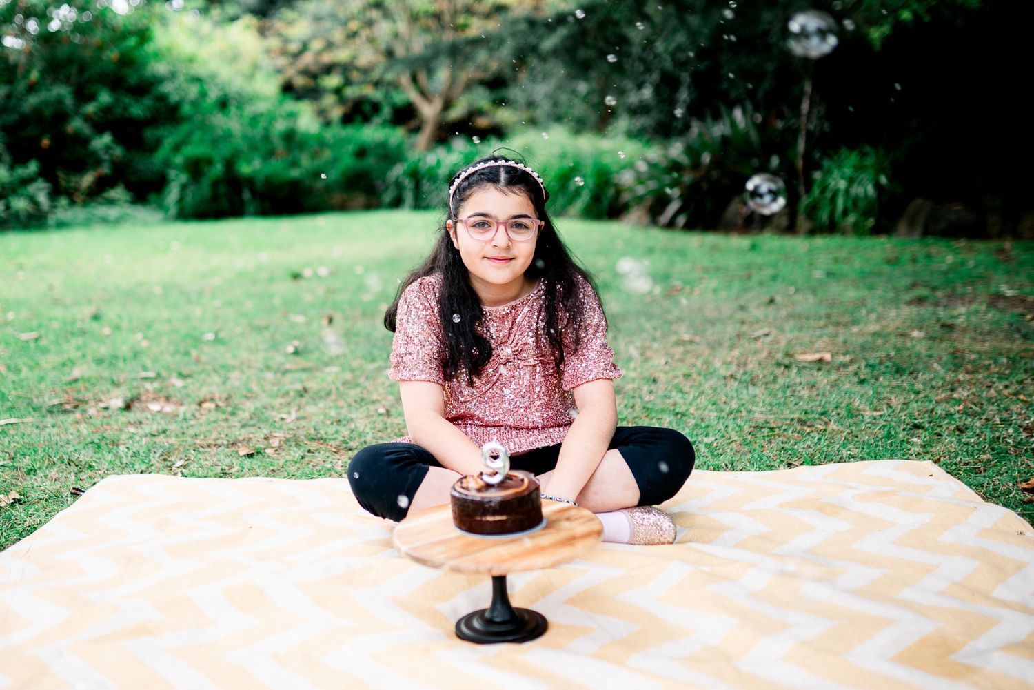 Birthday girl smiling in a family photoshoot in Melbourne's St Kilda Botanical Gardens with lush greeneries and a birthday cake and bubbles