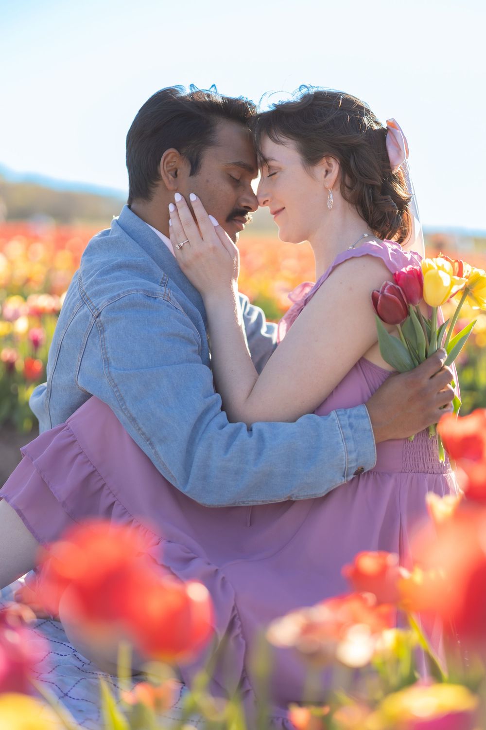 Couple embracing romantically in a vibrant tulip field, woman holding pink tulips, man in denim jacket, colorful flowers surrounding them.