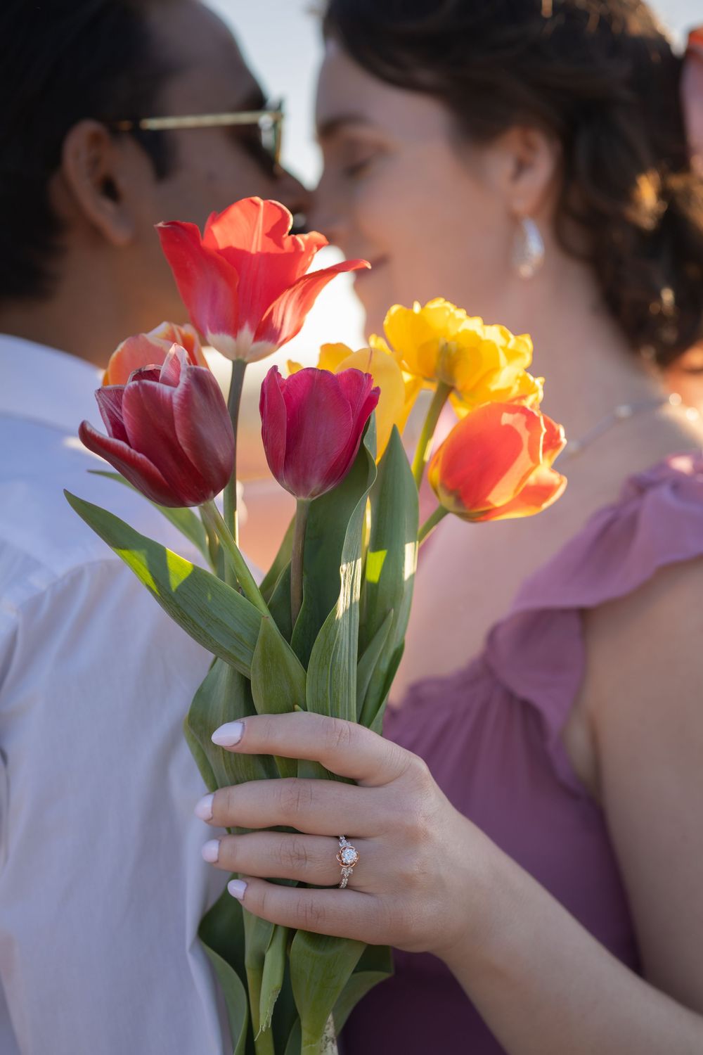 Couple sharing a romantic moment behind a vibrant bouquet of red and yellow tulips, woman wearing engagement ring.