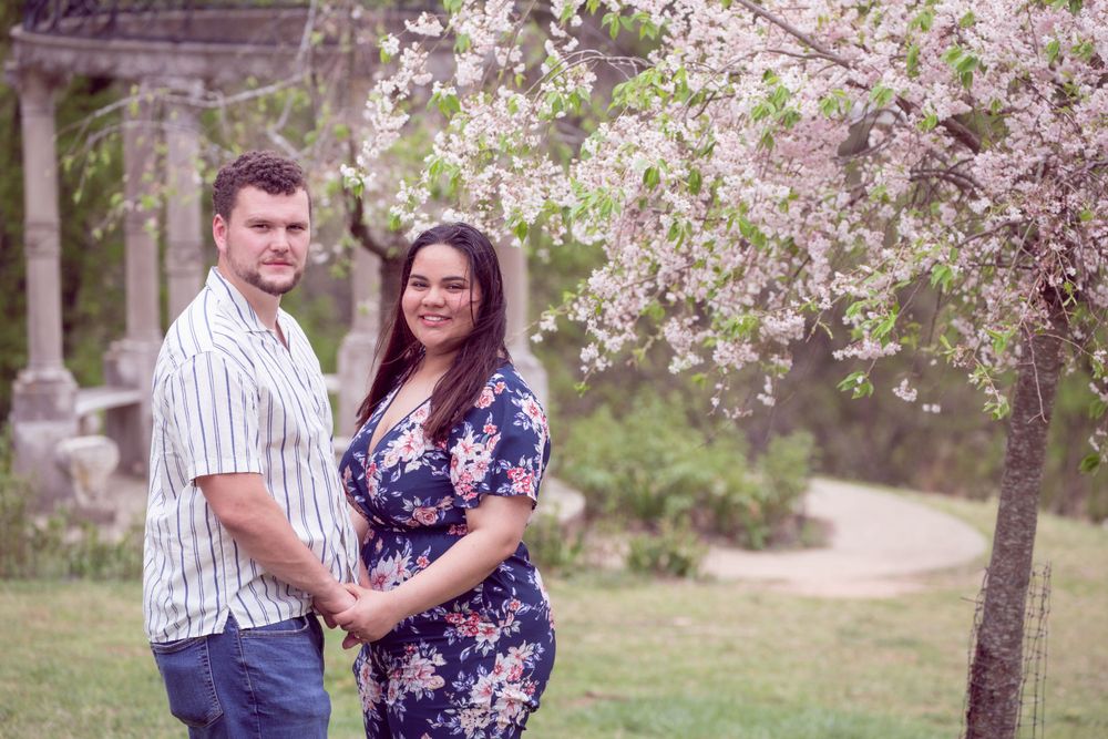 A romantic couple poses beneath flowering spring trees in a garden setting.