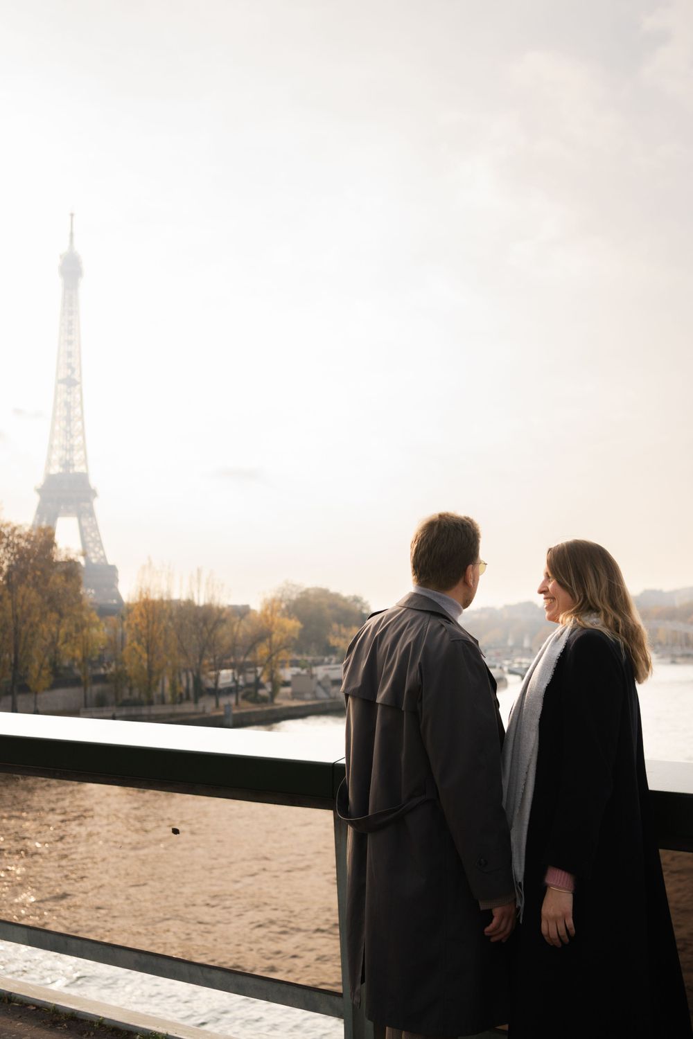 A couple in winter coats stands before the Eiffel Tower on an autumn day in Paris.