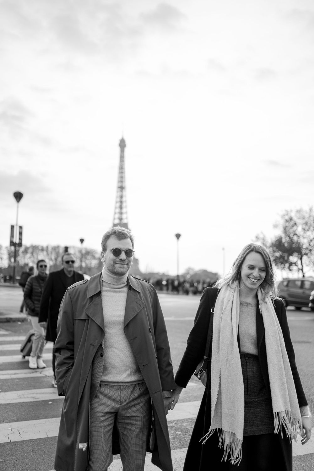 A couple walks hand in hand on a Paris street with the Eiffel Tower visible in the cloudy background.