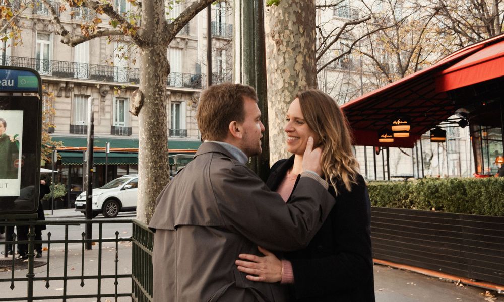 A couple embraces romantically on a Paris sidewalk near a red awning with trees and buildings in the background.