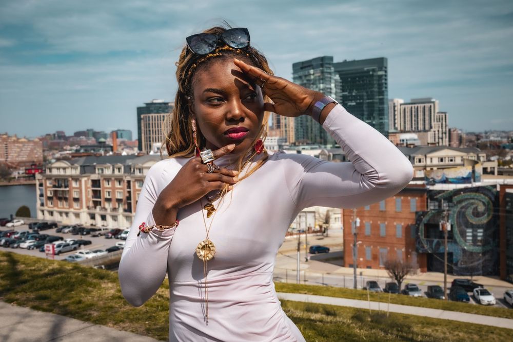 Stylish person in white long-sleeve top poses expressively against urban cityscape backdrop on sunny day.