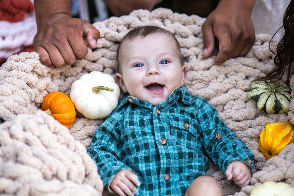 A baby wearing a teal plaid shirt lies on a cream knit blanket next to small decorative pumpkins.