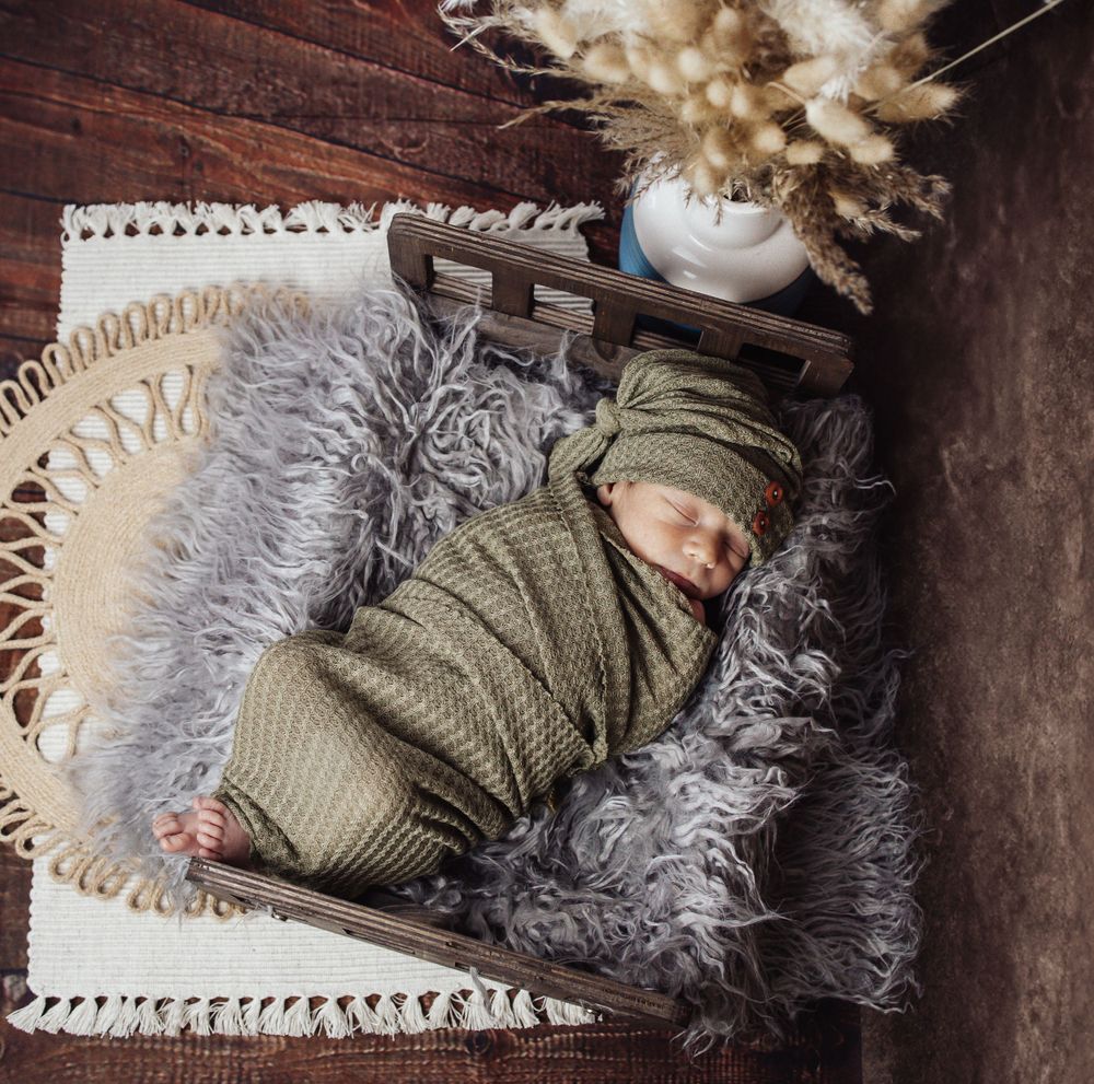 Sleeping newborn wrapped in olive green swaddle rests on textured gray fur blanket with rustic wooden backdrop and dried florals.