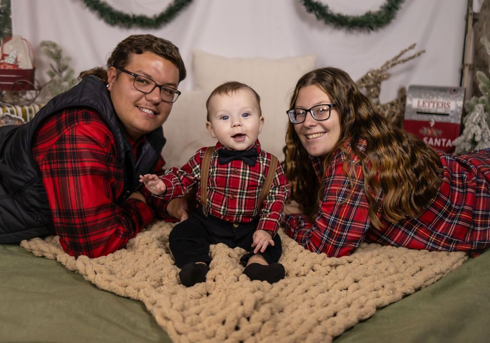 A family wearing matching red plaid outfits poses together on a beige knit blanket for holiday photos.