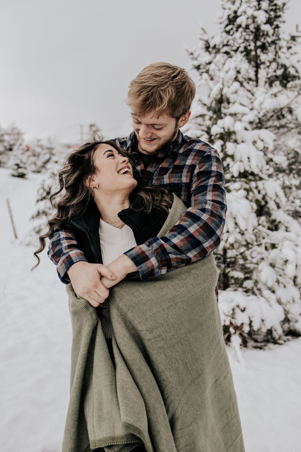 A couple wrapped in a cozy olive blanket share joyful moments together in a snowy winter setting.