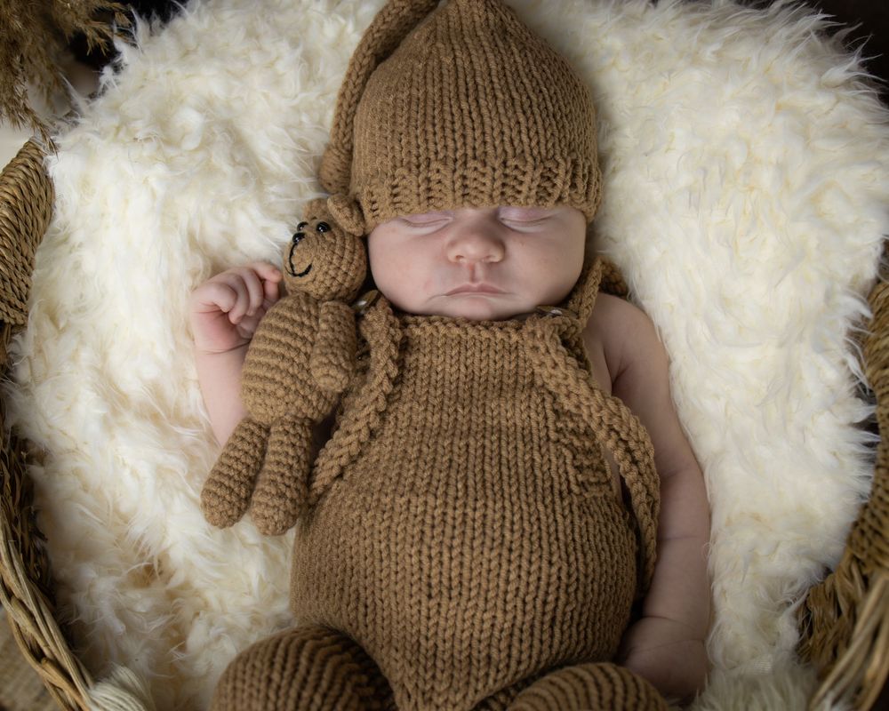 Newborn baby wearing brown knitted outfit and hat lying on soft white fur blanket.