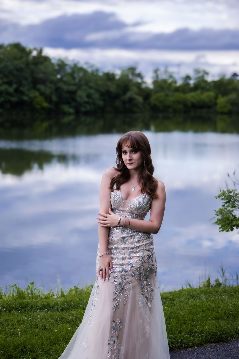 Figure in an elegant beaded gown poses by a serene lake during twilight with trees reflecting in the calm water.