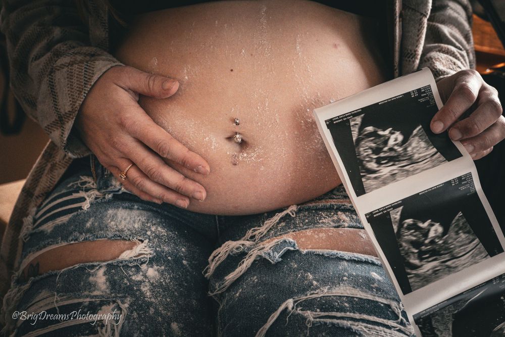 A pregnant woman in ripped jeans holds ultrasound photos while touching her baby bump.