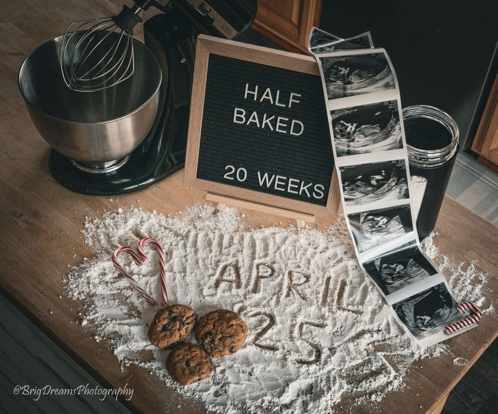 Pregnancy announcement featuring 'Half Baked 20 Weeks' sign with ultrasound photos and date written in flour on dark surface.