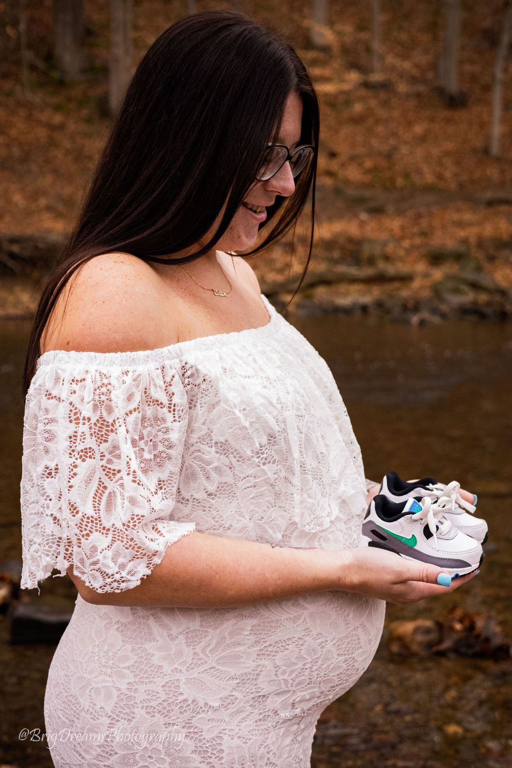 An expectant mother in lace dress holding baby shoes by a stream.