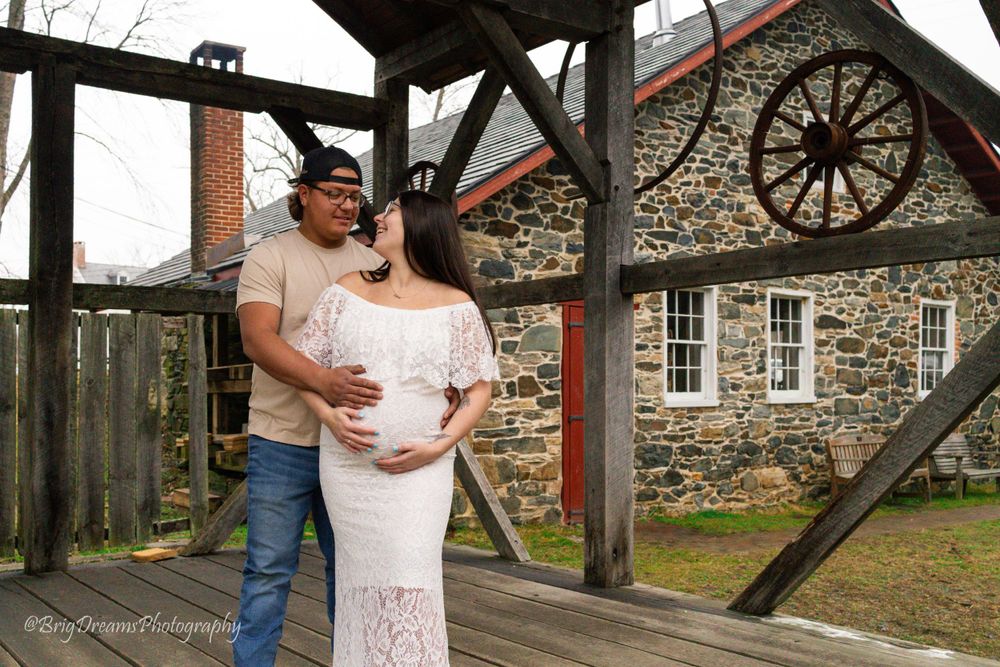 A couple poses together on a rustic wooden deck with a stone building and wagon wheel in the background.