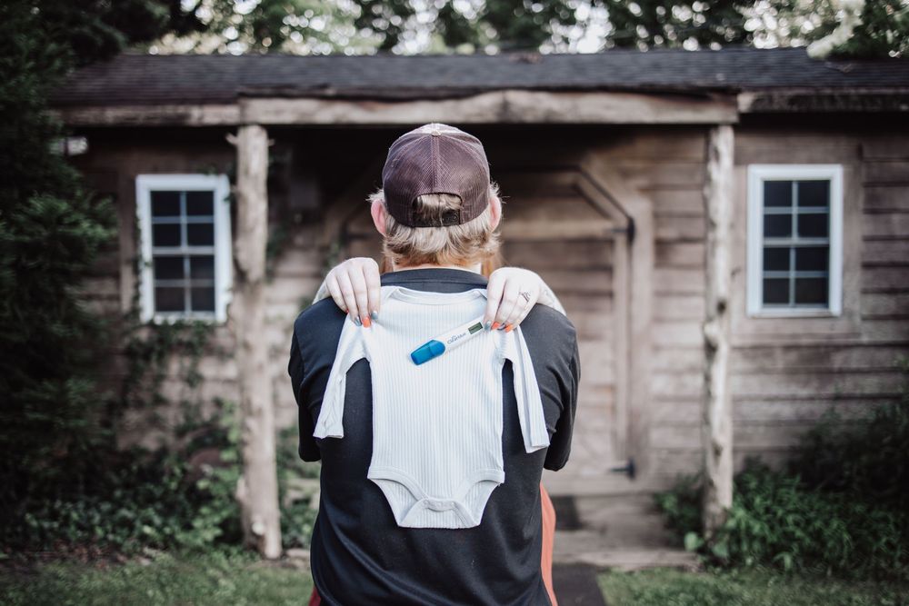 A person showing a baby onesie against a rustic wooden cabin background in dim lighting.