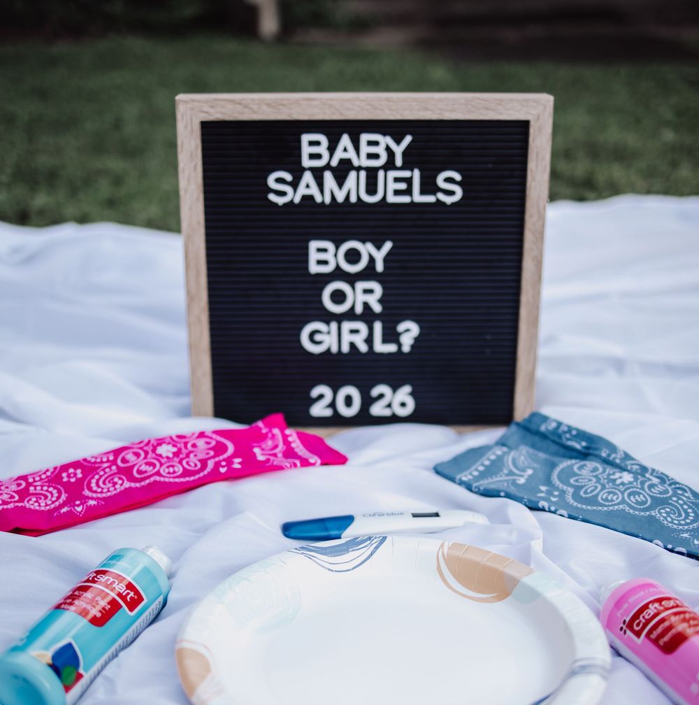 Gender reveal announcement board with 'Baby Samuels' and pink and blue bandanas on white blanket.
