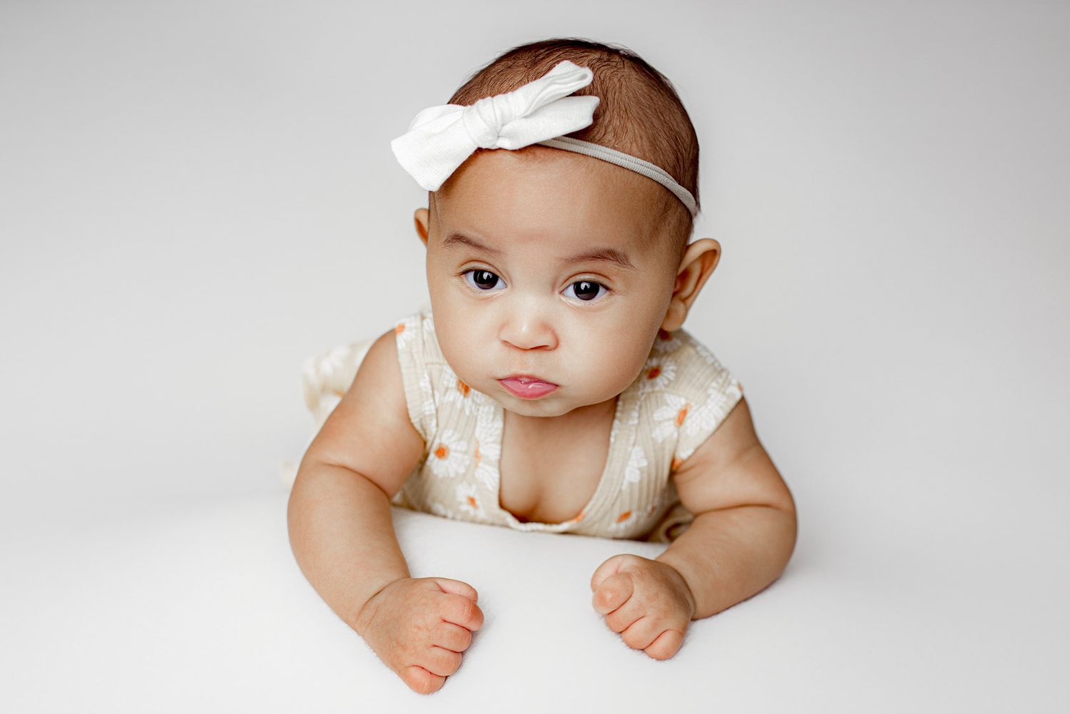 Baby wearing white lace outfit and bow headband lies on white backdrop during professional photo session.