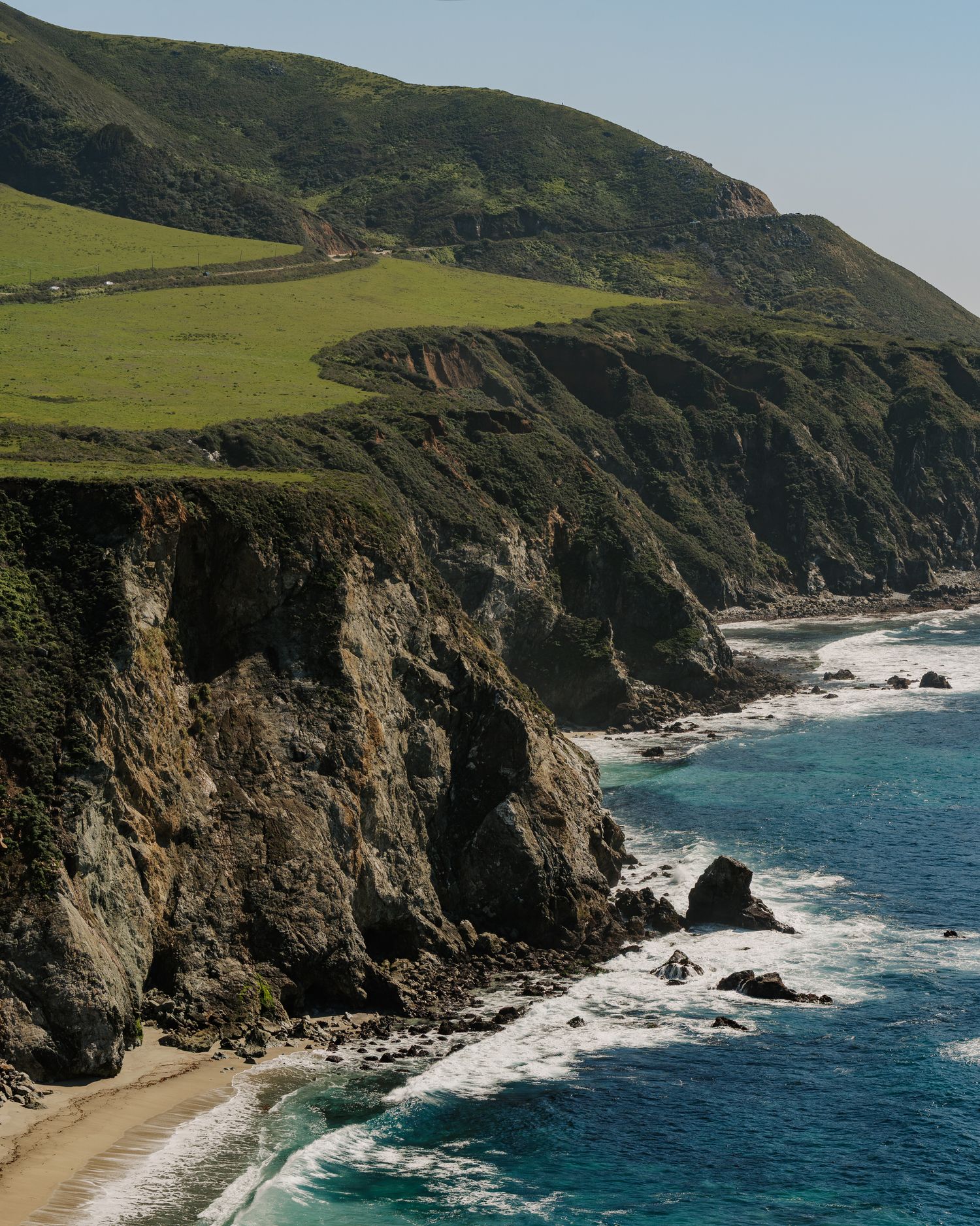 Rocky coastal cliffs meet turquoise waters along Big Sur's dramatic coastline with green rolling hills above.