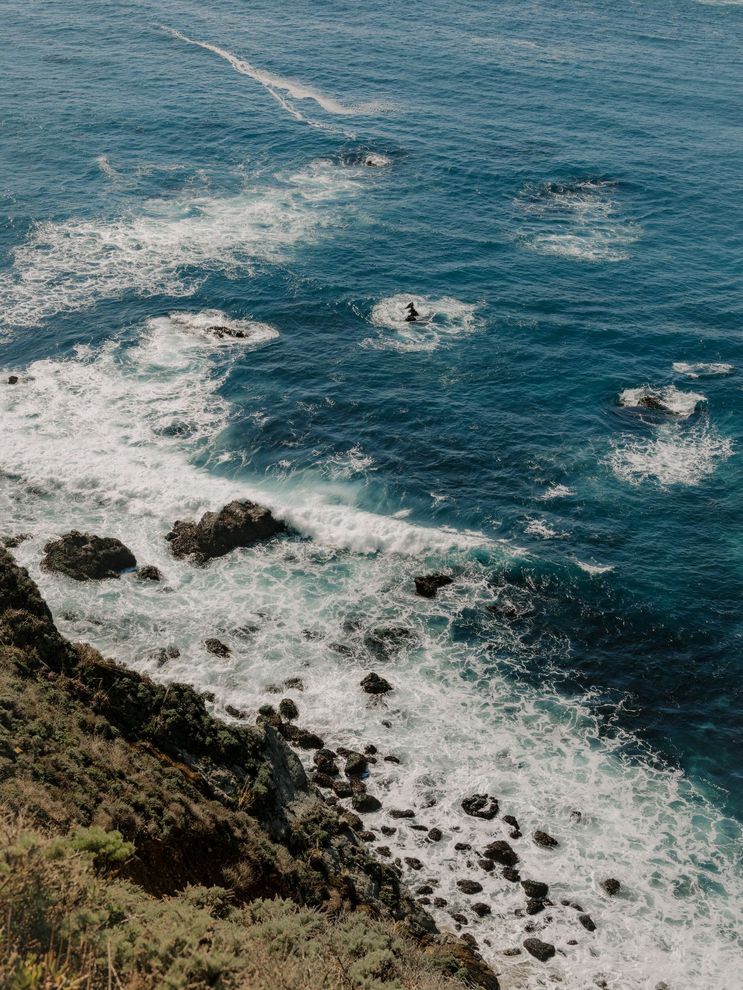 Aerial view of rocky coastline with crashing waves against jagged rocks in deep blue ocean waters.