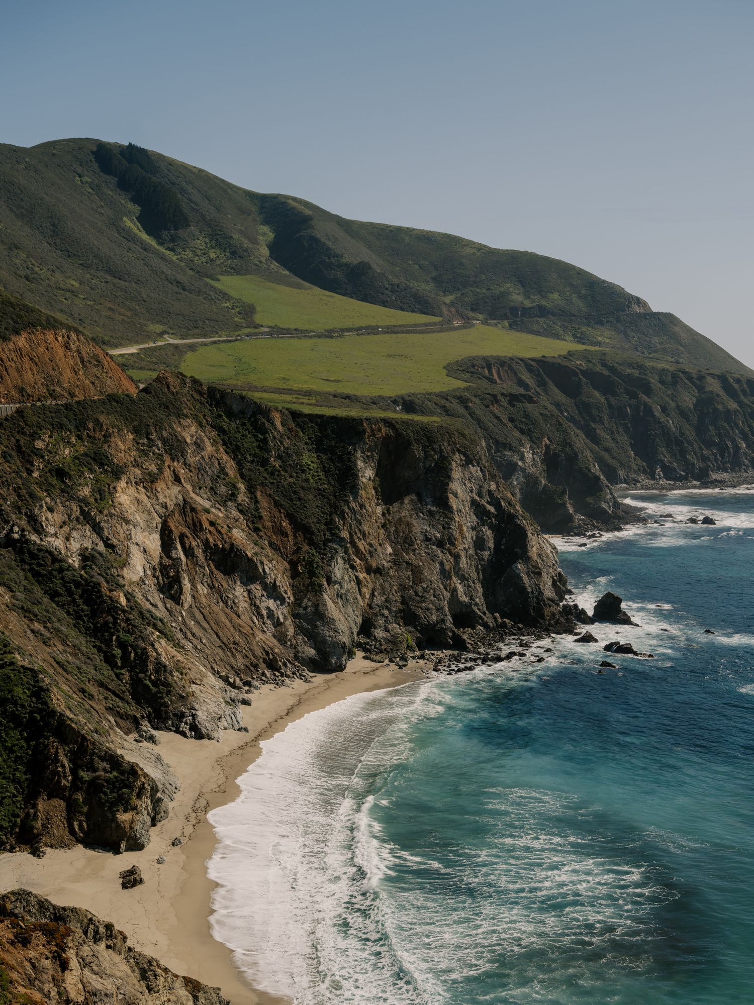 Rugged coastal cliffs and turquoise waters along the dramatic Big Sur coastline with waves crashing on a secluded beach.