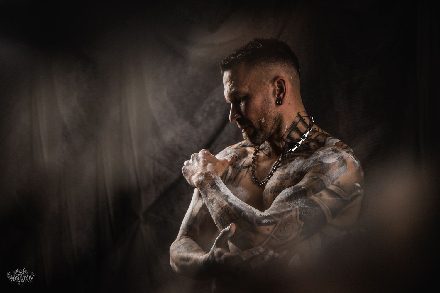 Moody portrait of tattooed figure emerging from smoke against dark background.
