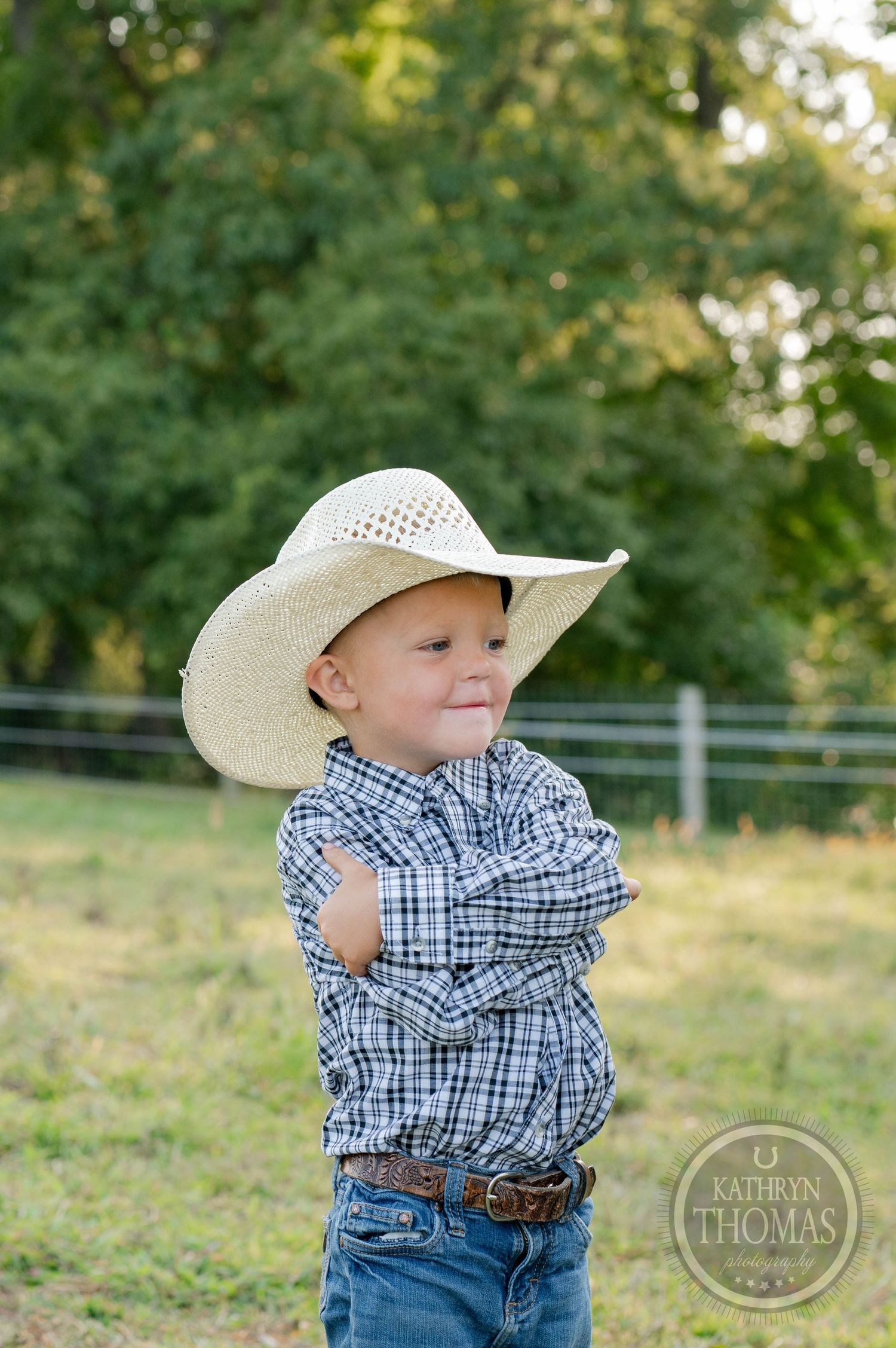 Young ranch hand in cowboy hat and plaid shirt poses against rustic farm background.
