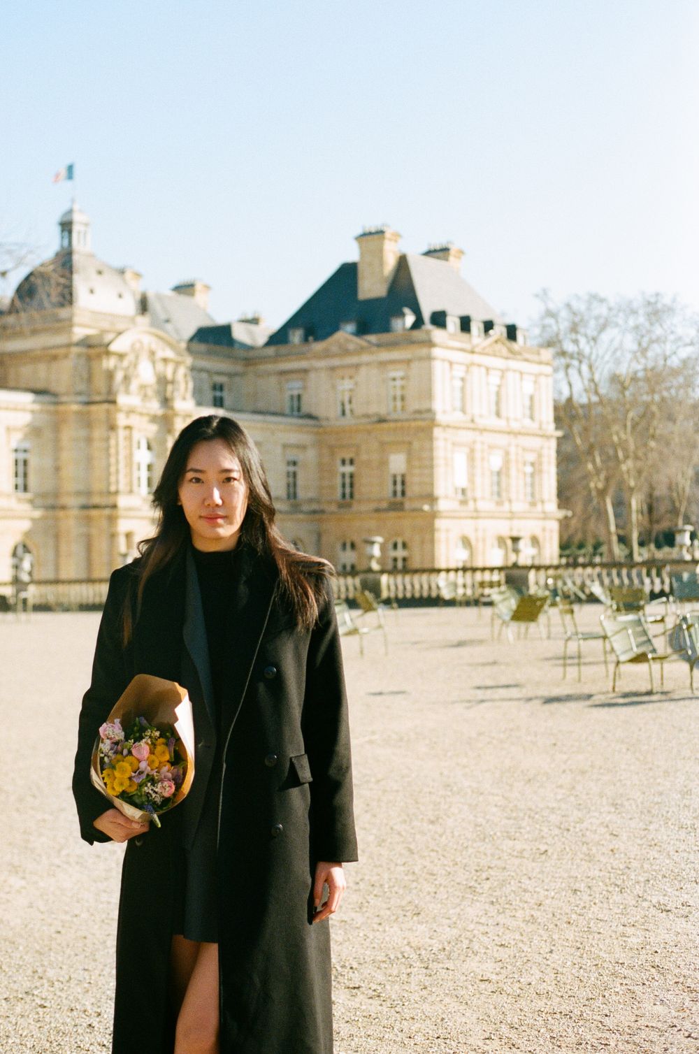 Standing in front of Luxembourg Palace on a sunny day, wearing a black coat and holding flowers in the garden.