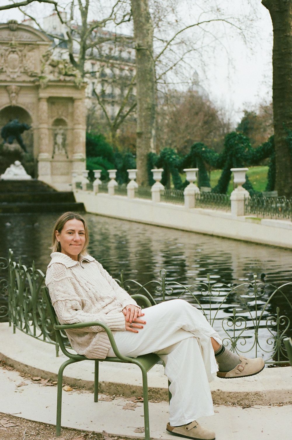 A person relaxes in a green chair near a reflecting pool in a Parisian garden with classical architecture.